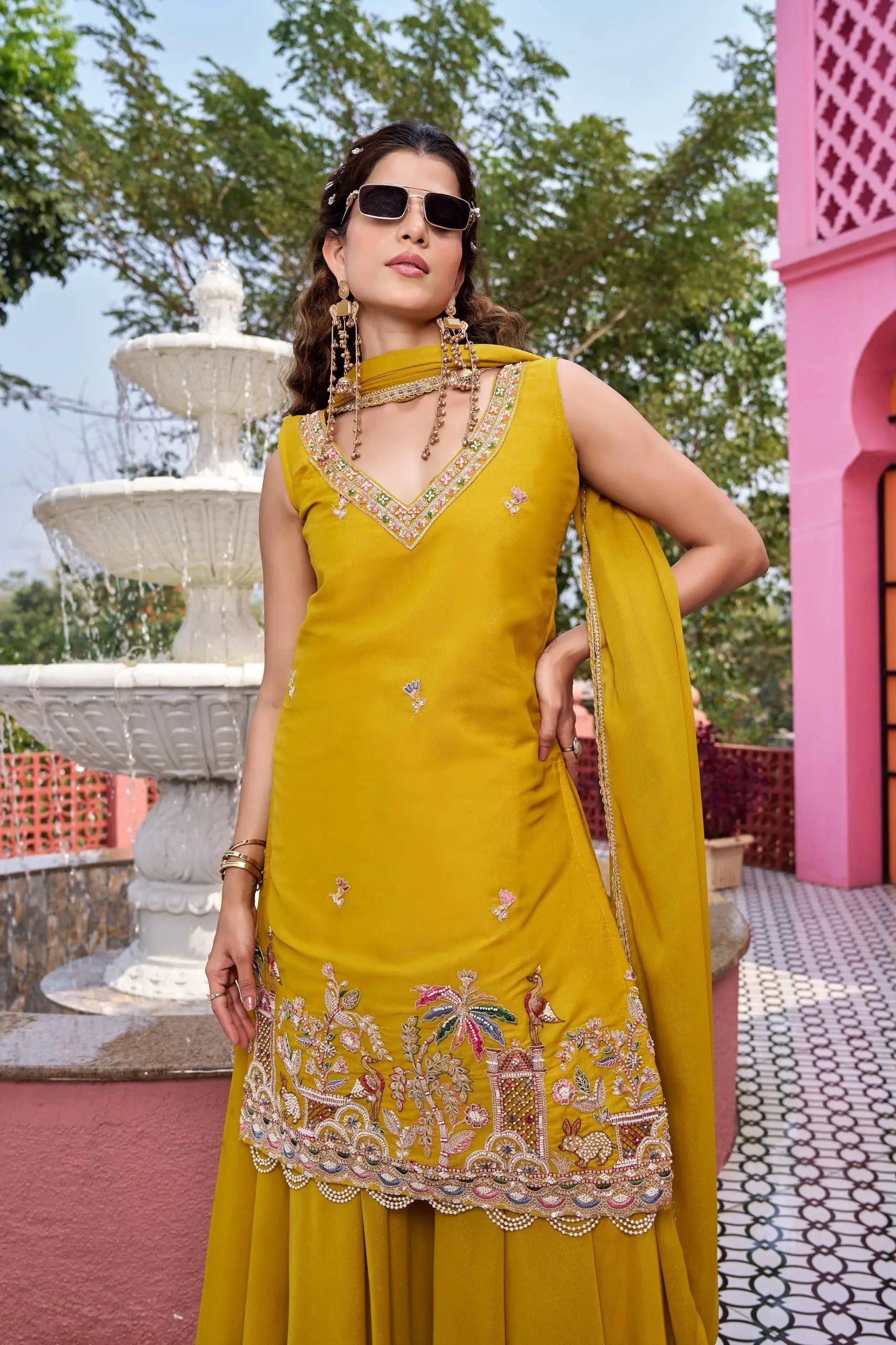 Woman in a yellow traditional outfit with intricate designs, standing outdoors near a fountain and pink building.